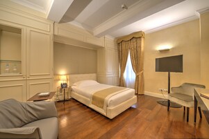 Habitación de hotel con cama, sofá, escritorio, lámpara, televisor y ventana con cortinas.