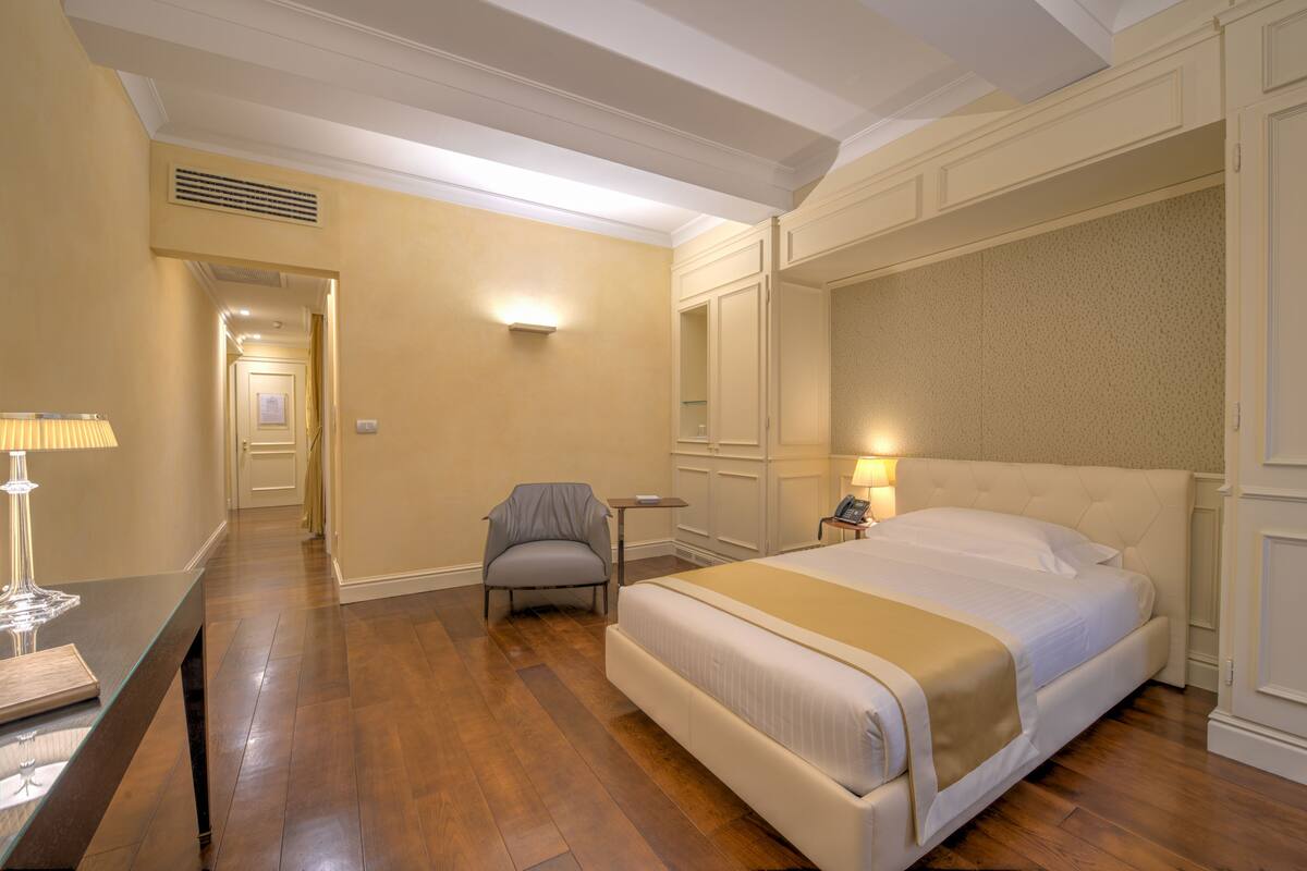 A hotel room with a large bed, a chair, a desk with a lamp, and a nightstand with a phone.