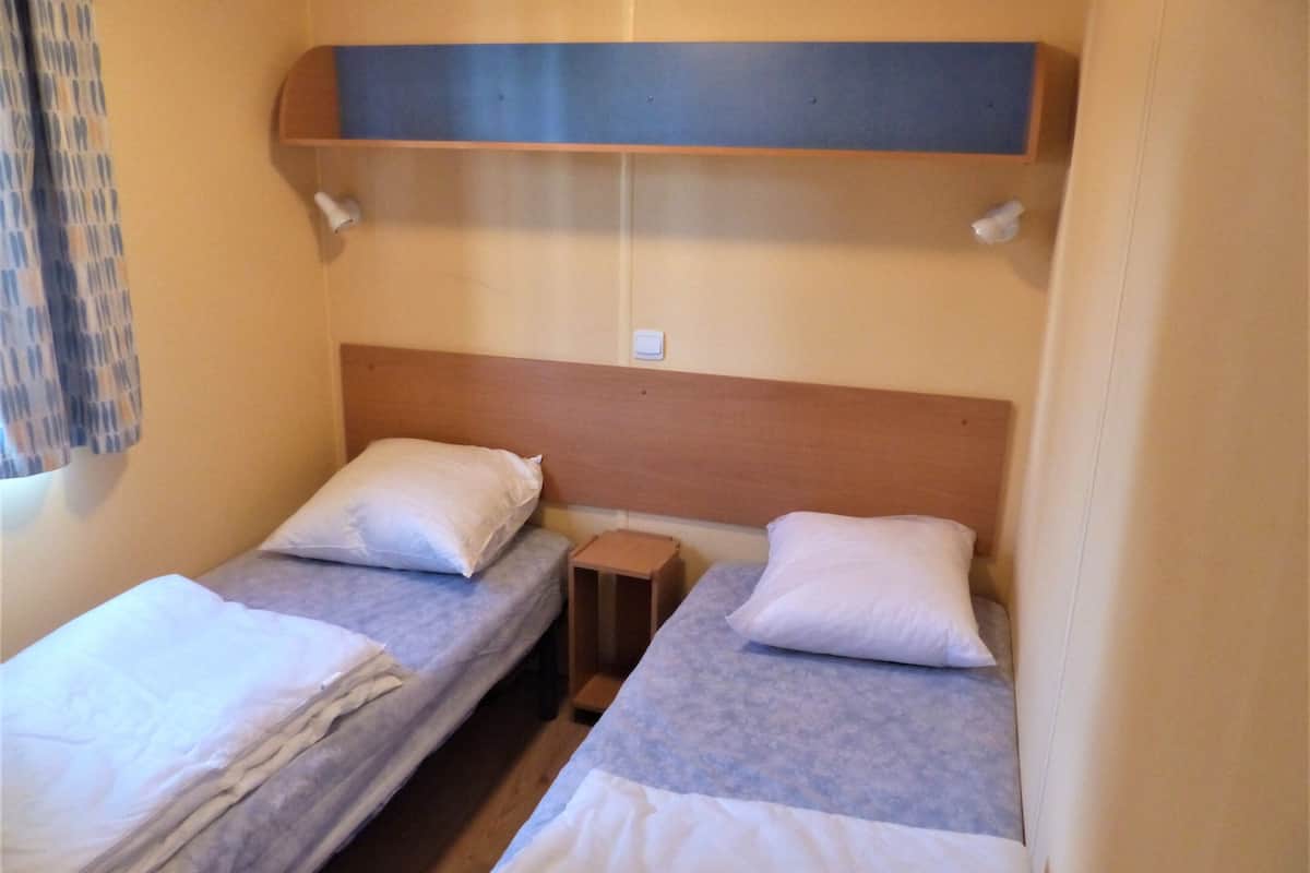 A small, compact room with two single beds, each with a wooden headboard and white bedding. There is a small wooden nightstand between the beds, and a shelf above the beds.