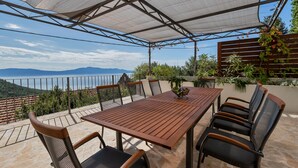 Un patio al aire libre con mesa y sillas de madera, zona cubierta con vistas al mar y las montañas, y plantas en macetas.