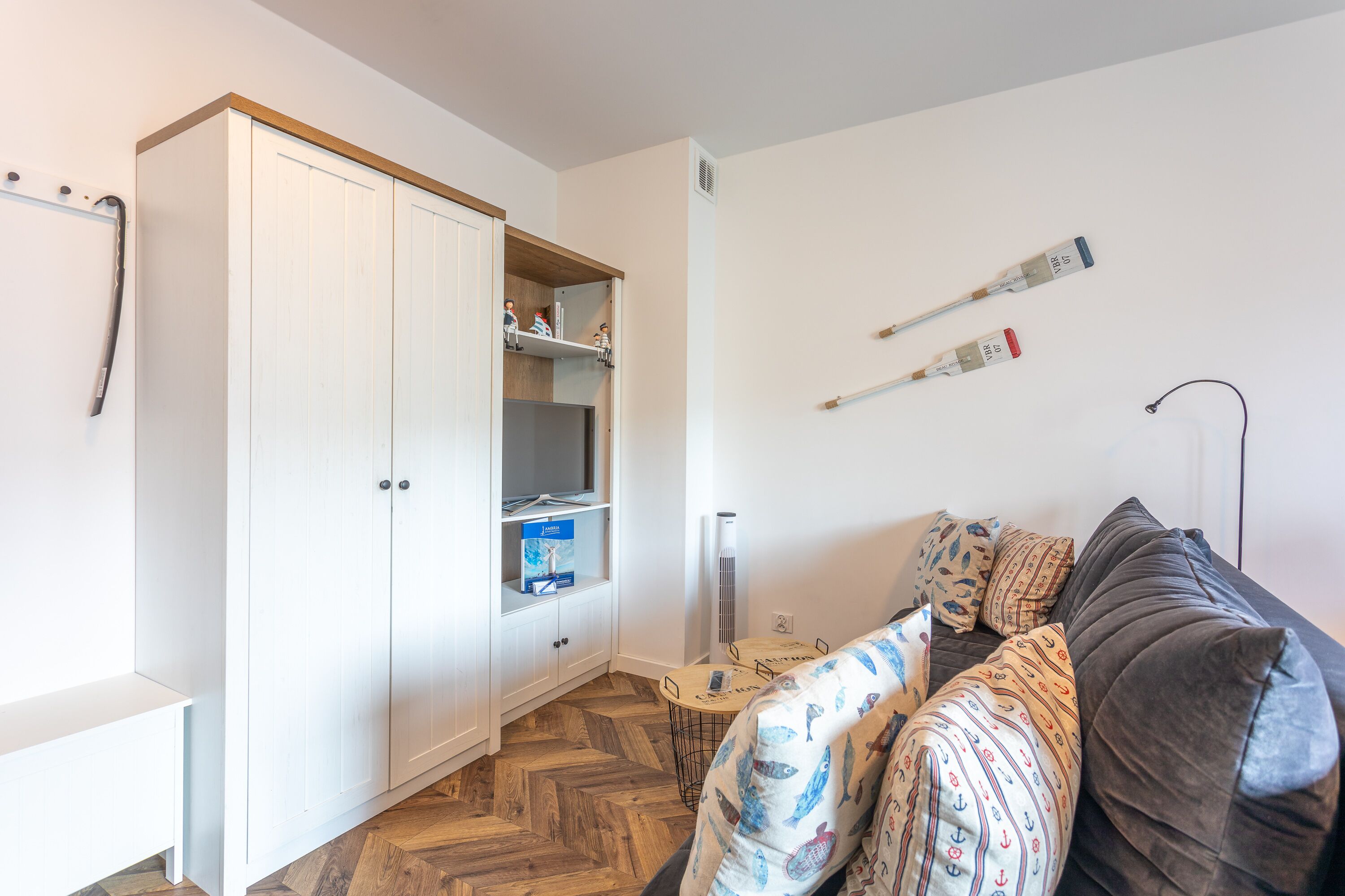 Una habitación con un armario blanco, un sofá oscuro, suelo con estampado de espiga y una pequeña mesa con una silla.