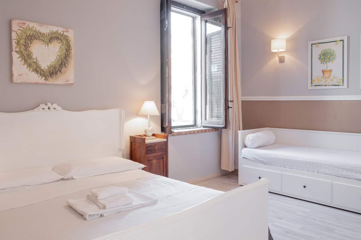 A bedroom with a bed, a nightstand, a bedside table, a window, and a wall with a heart-shaped decoration and a framed picture.
