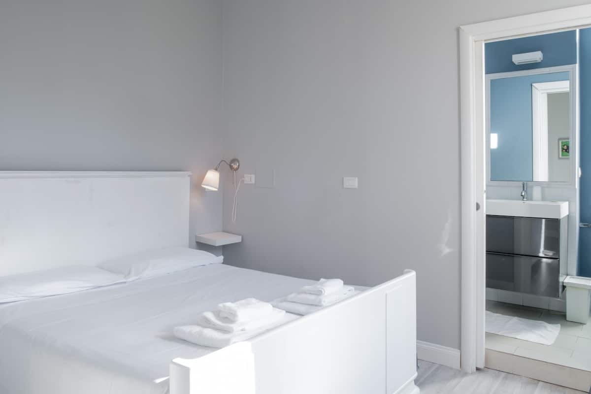 A modern hotel room with a neatly made bed, a bedside lamp, and a bathroom with a mirror and sink.