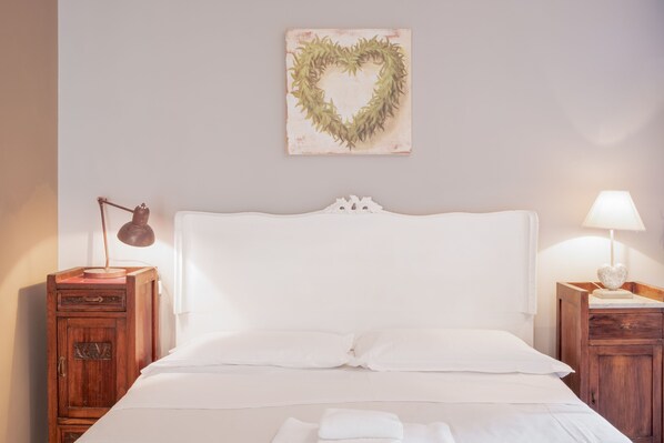 Una cama bien hecha con sábanas blancas, un adorno en forma de corazón en la pared y dos mesitas de noche con lámparas.