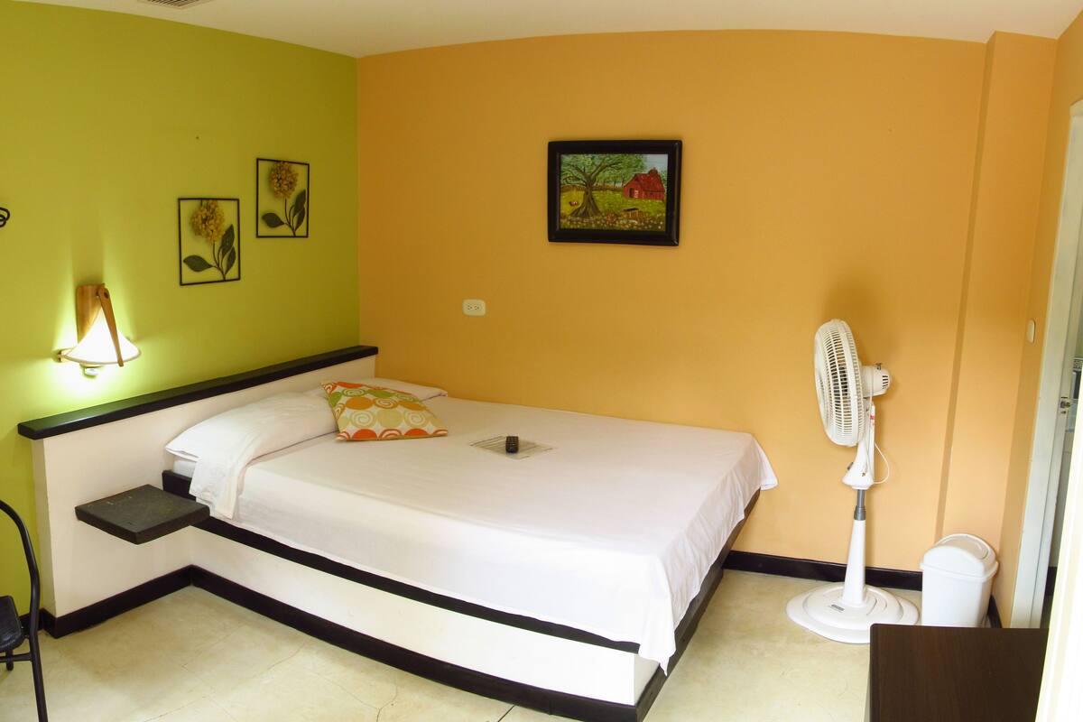 Un lit simple avec un matelas blanc et une tête de lit en bois, une petite table avec deux chaises, un ventilateur fixé au mur et deux tableaux encadrés accrochés au mur.