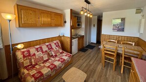 A cozy living area with a wooden sofa, a dining table with chairs, a kitchen with wooden cabinets, and a hanging light fixture. - 2-room apartment with parking in Les Saisies, 4 people, balcony, fully equipped (Les Saisies)