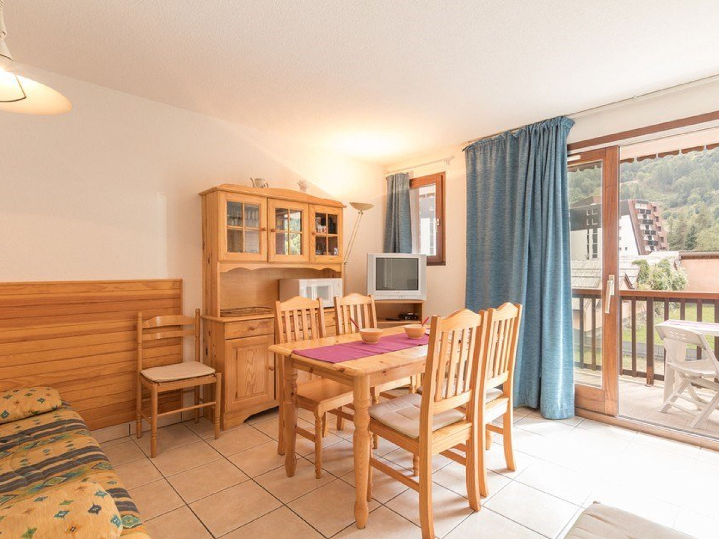 A cozy room with a wooden dining table, chairs, a cabinet, a television, and a balcony with a view of the landscape.