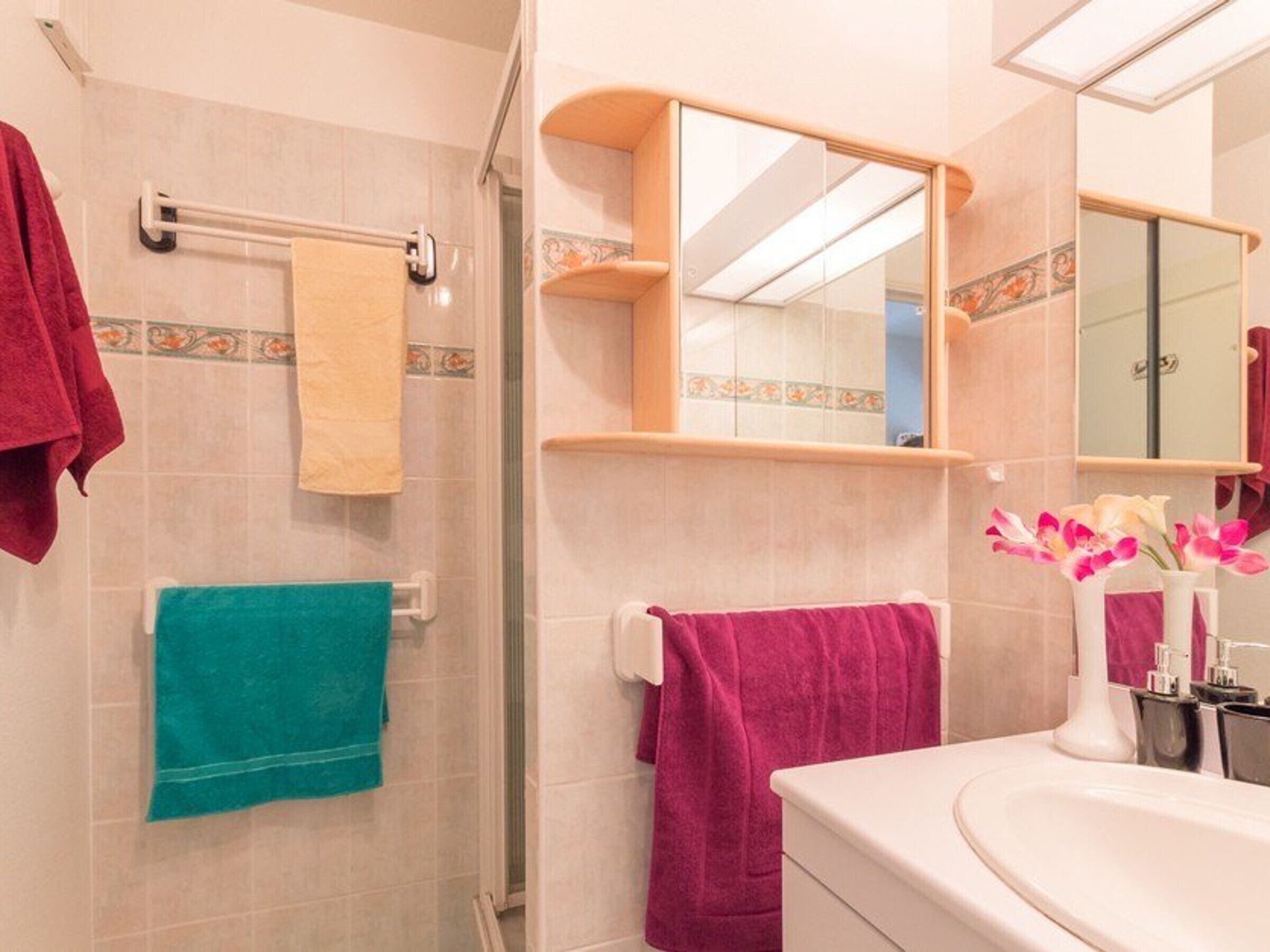 Une salle de bain avec une douche, un lavabo et un miroir. Des serviettes sont accrochées au mur et un vase rempli de fleurs est posé sur le lavabo.