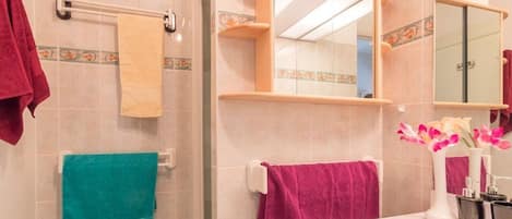 A bathroom with a shower, a sink, and a mirror. There are towels hanging on the wall and a vase with flowers on the sink.
