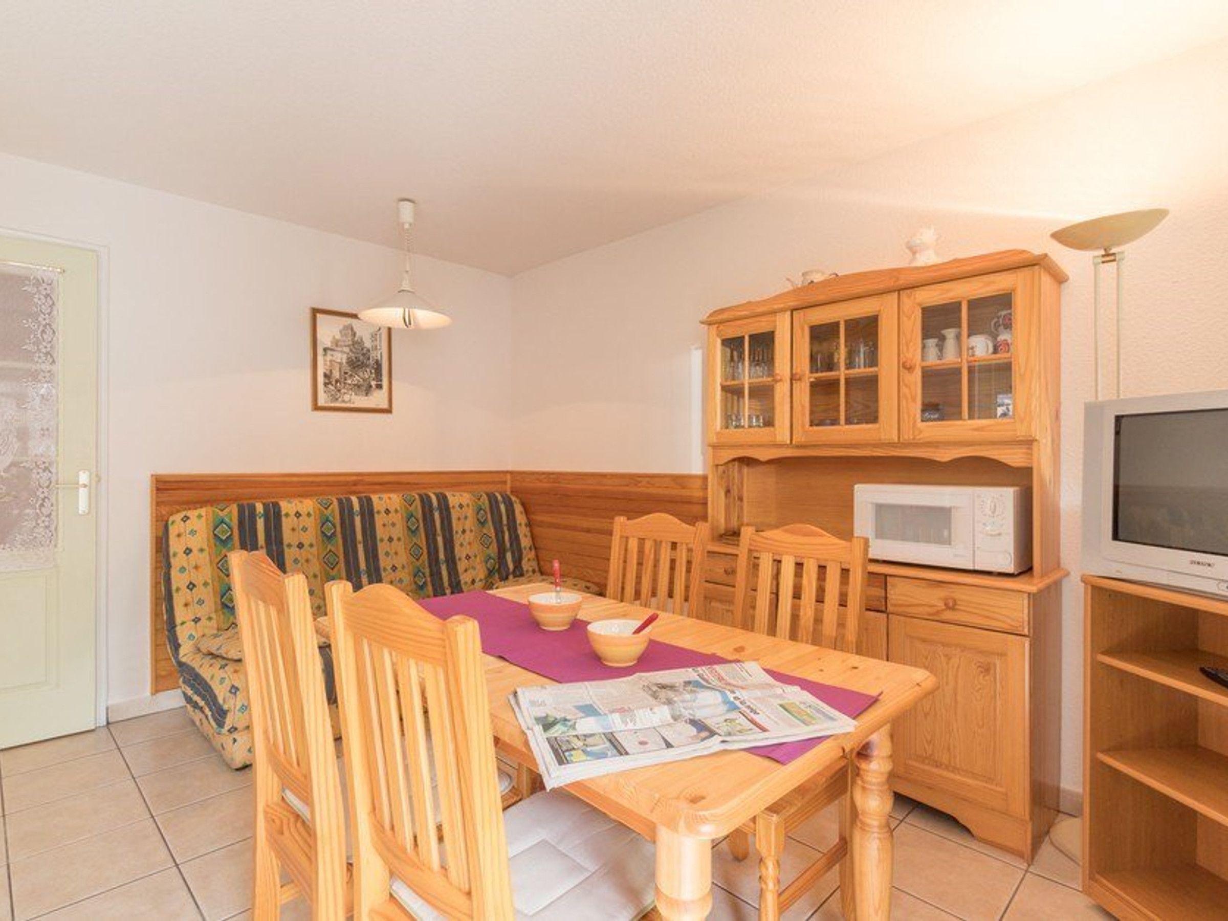 A cozy living room with a wooden sofa, a dining table with chairs, a cabinet with glass doors, a microwave, and a television.