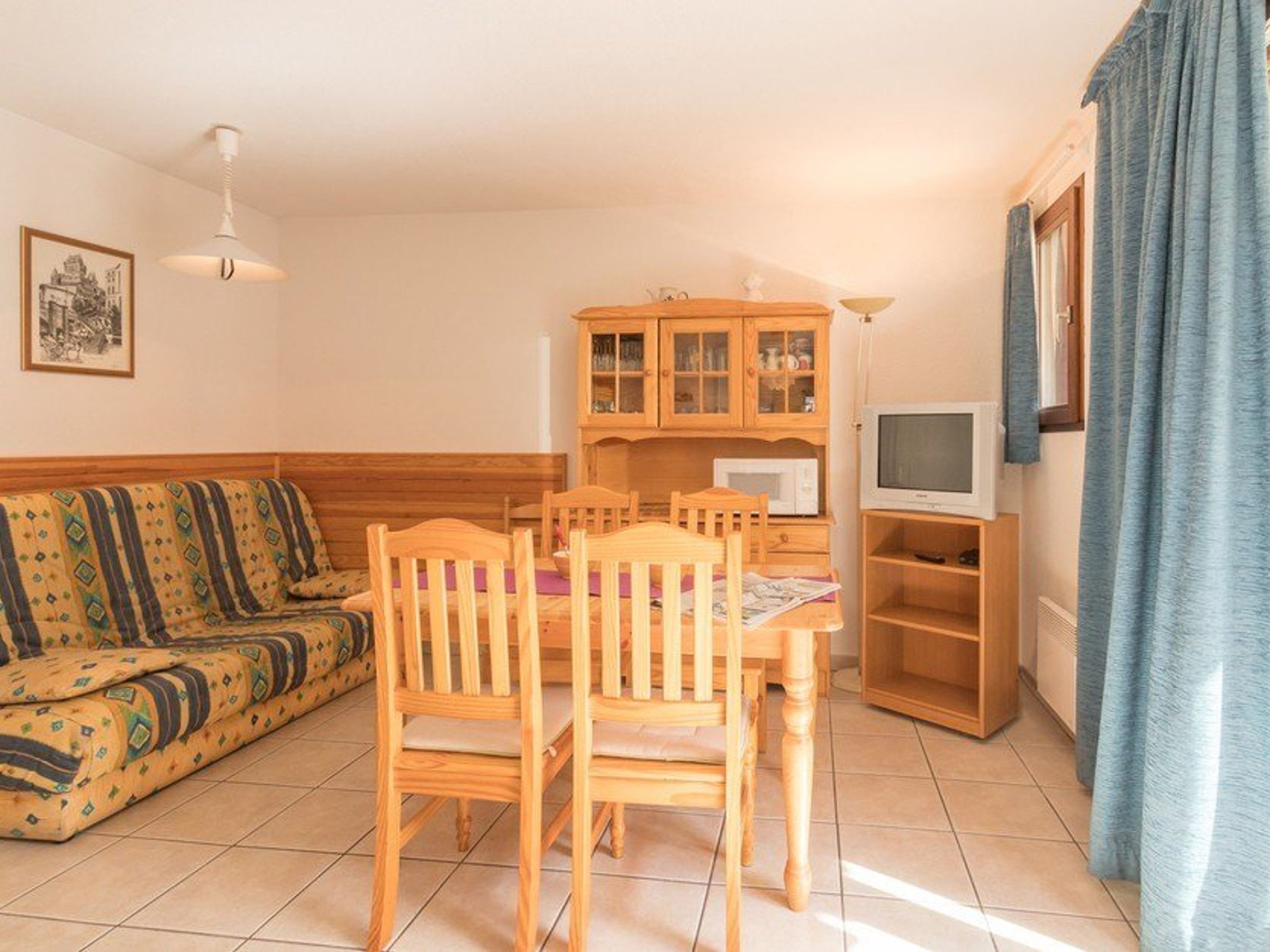 En stue med sofa, træstole, spisebord, fjernsyn og et køkkenområde med skabe og en mikroovn.