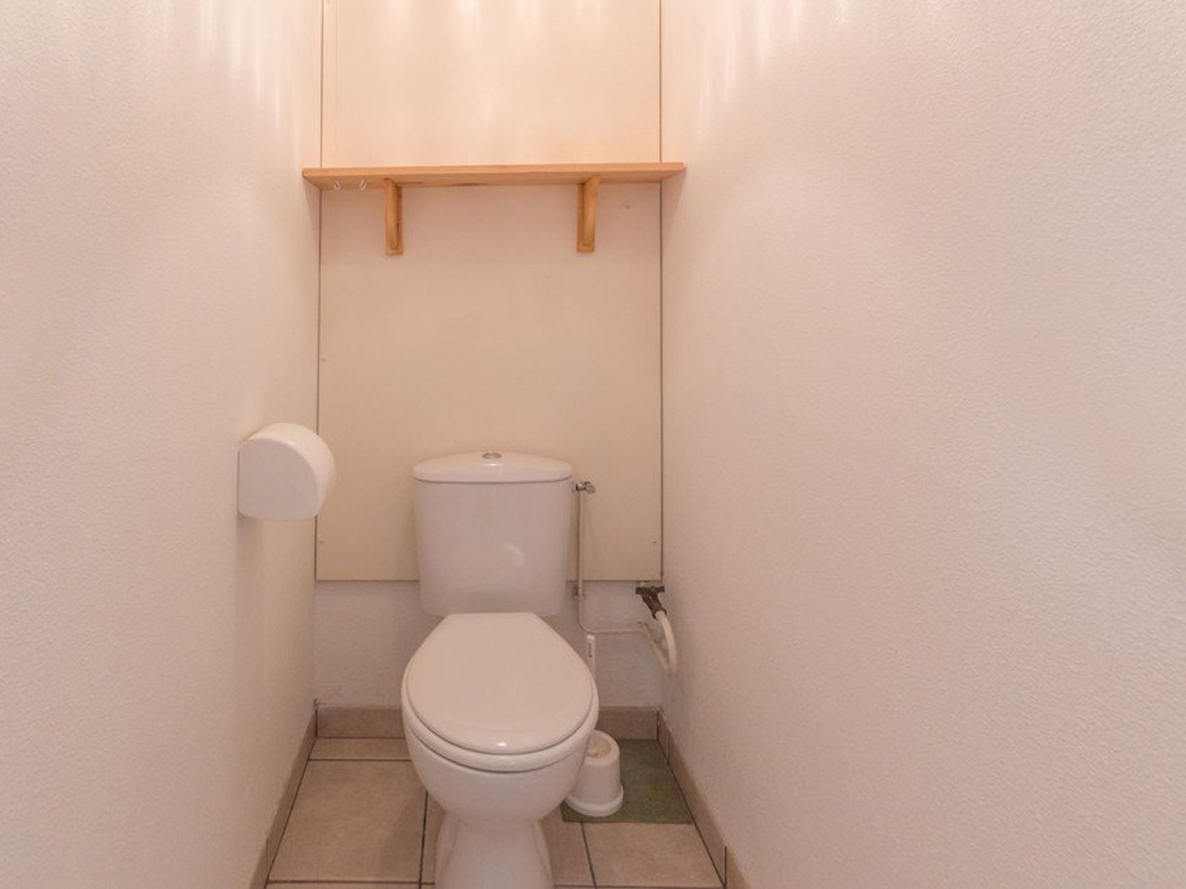 Een kleine, schone badkamer met een toilet, een toiletrolhouder en een plank boven het toilet.