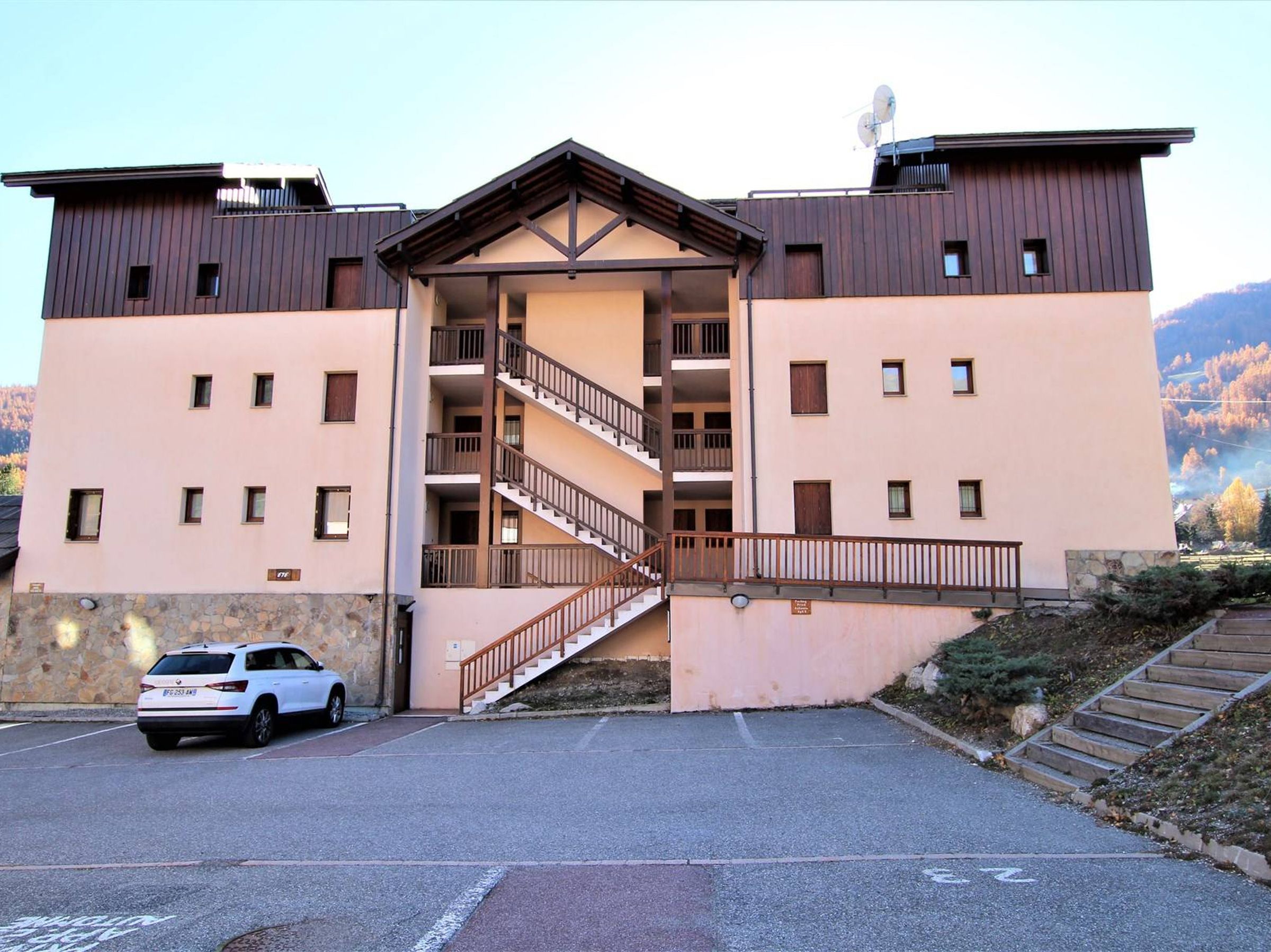 A multi-story building with a central staircase, a car parked in front, and a stone wall on the left.