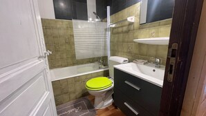 A compact bathroom with a bathtub, a toilet with a green seat, a sink, and a mirror above the sink. - Cozy studio with south-facing balcony and parking (Châtel)