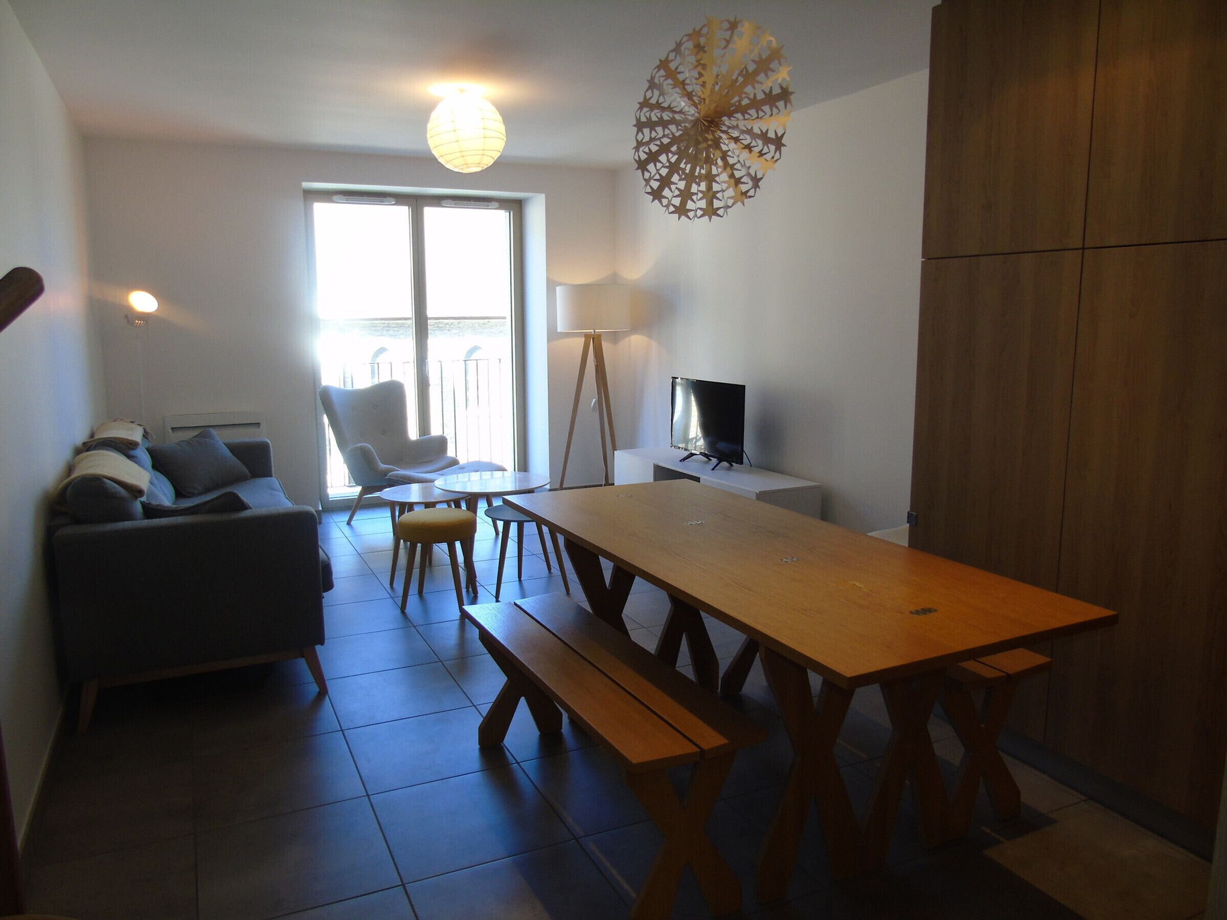 A modern living room with a sofa, a coffee table, a dining table with benches, a TV, and a decorative light fixture.