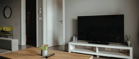 A modern living room with a wooden coffee table, a flat-screen TV on a white stand, and a door leading to another room.