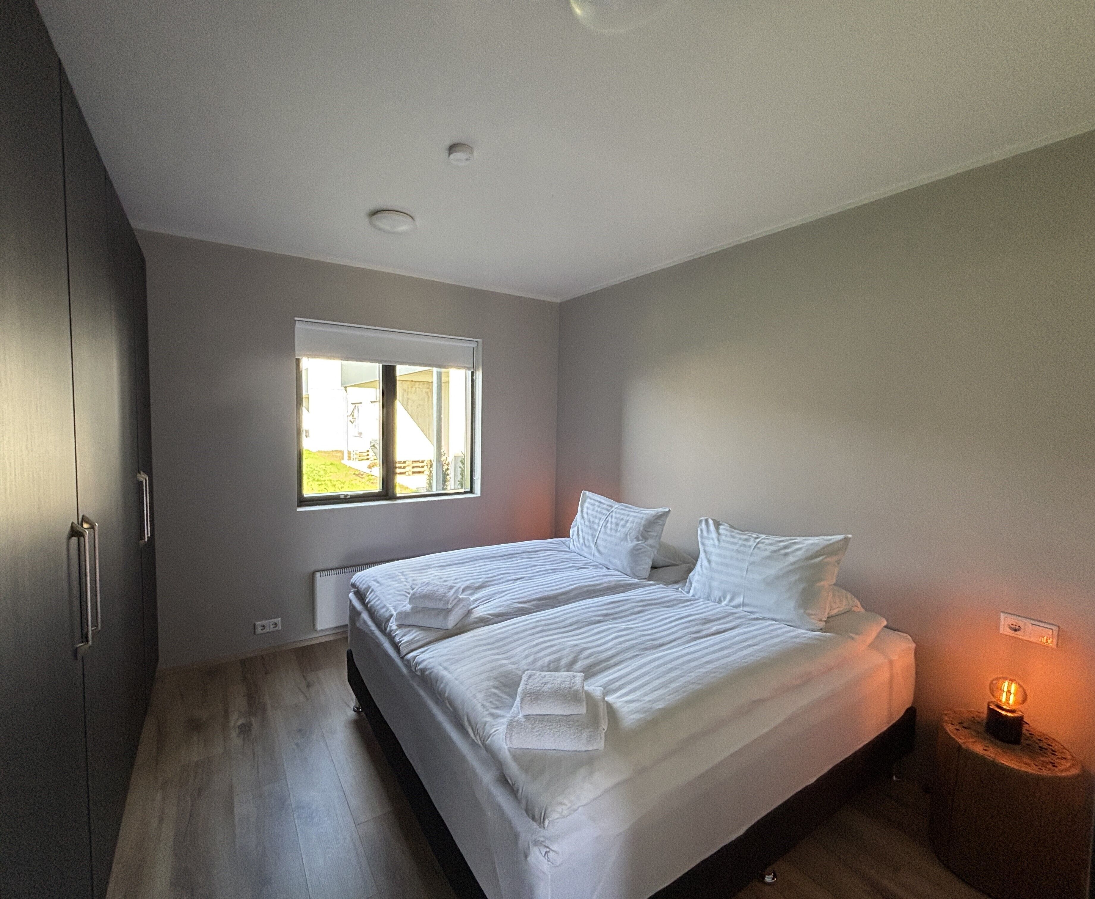 A bedroom with a bed, a window, a bedside table with a lamp, and a wardrobe.