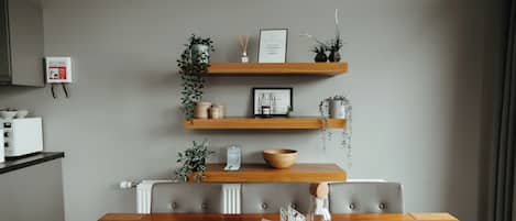 Un comedor moderno con una mesa de madera, dos estantes de madera con plantas y adornos, un detector de incendios blanco instalado en la pared y una silla tapizada de color gris.