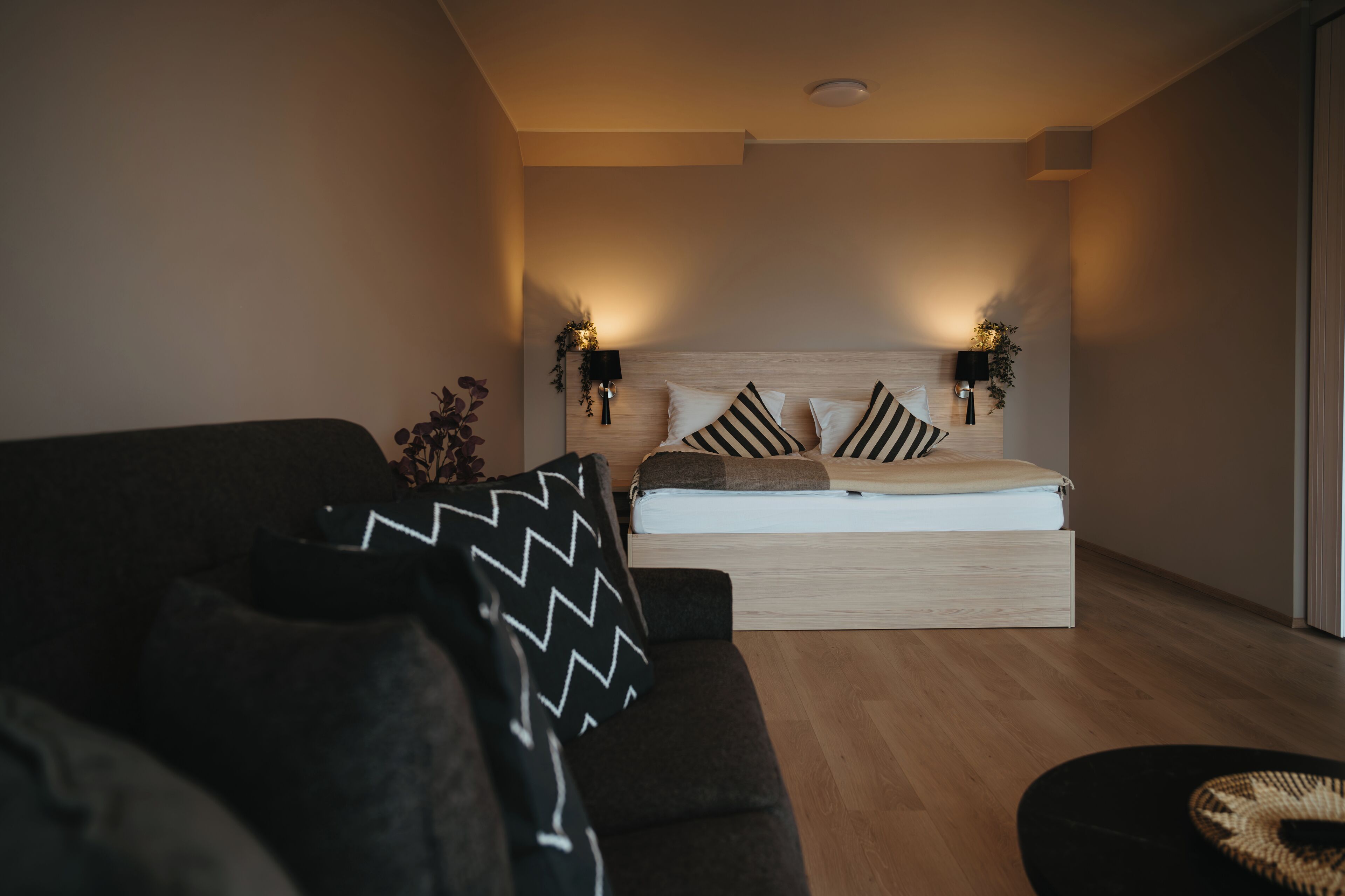 A modern bedroom with a large bed, two bedside lamps, a dark sofa, and a small round table.
