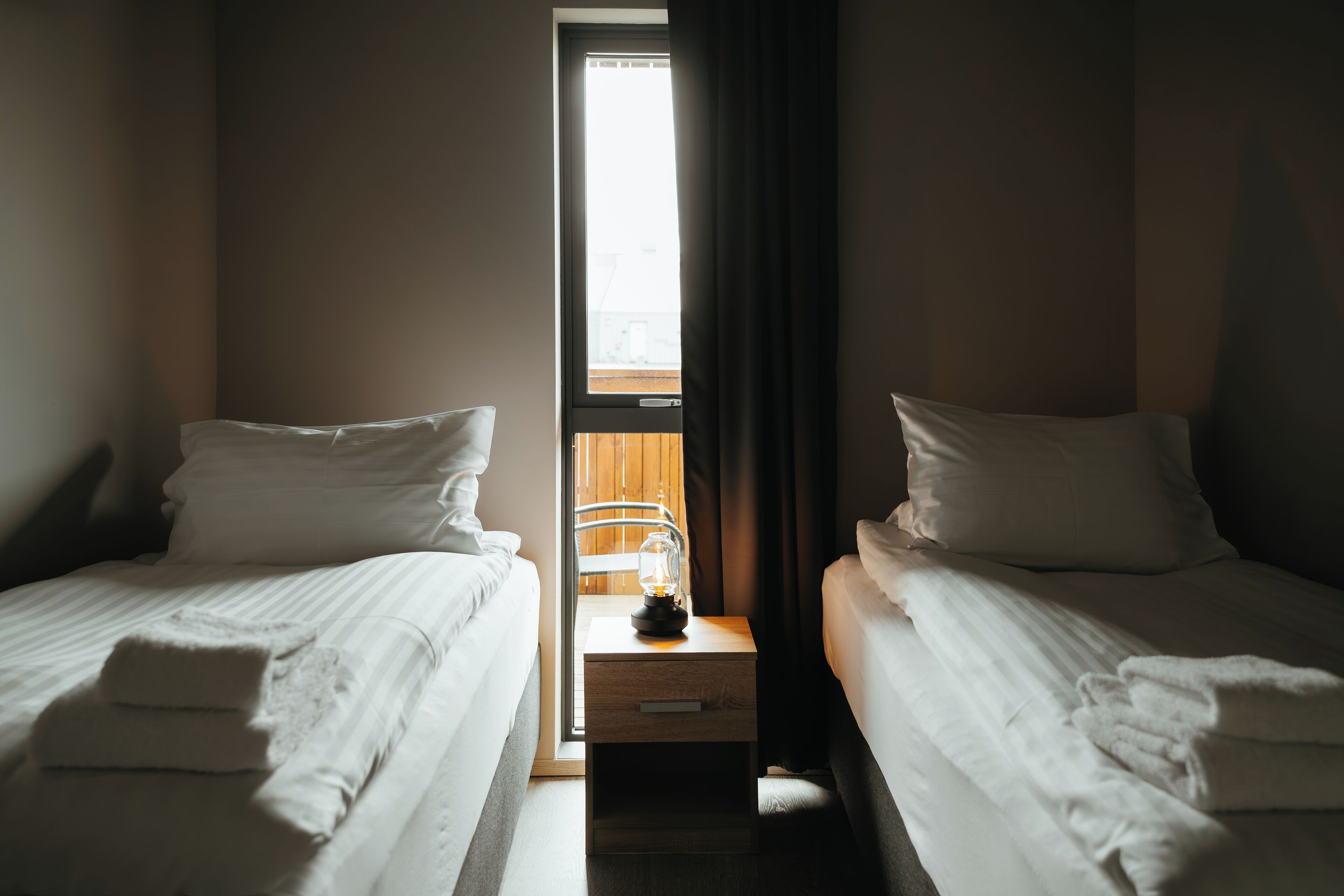 A hotel room with two single beds, a small table with a lamp, and a window with curtains.
