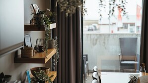 A modern interior with a wooden shelf holding decorative items and plants, a dining table with chairs, and a view of the outside through large windows. - Vík Apartments (Vik I Myrdal)