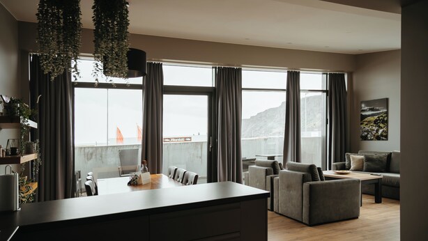A modern living room with a large window, a sofa, a coffee table, and a dining area. - Vík Apartments (Vik I Myrdal)
