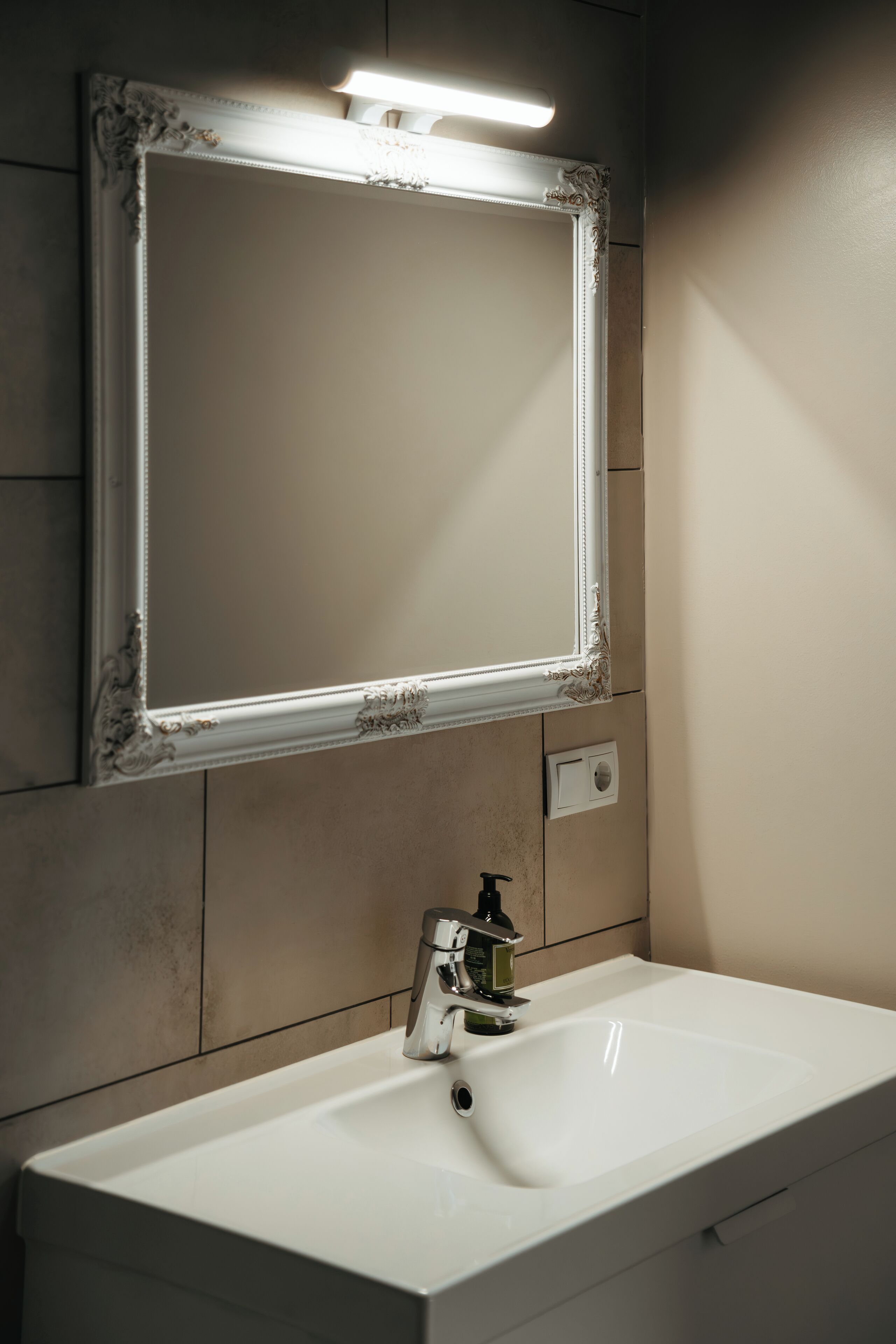 A bathroom with a white sink, a mirror with ornate framing, and a wall-mounted light fixture.