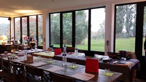 A dining area with large windows, tables set with water bottles, papers, and laptops. - Chambres d'hotes Maison Prairie Bonheur (Magny-les-Hameaux)