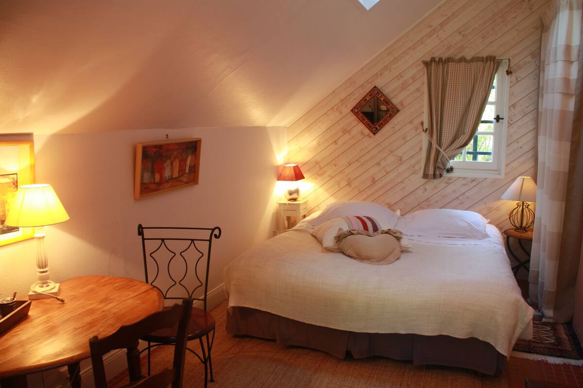 Une chambre confortable avec un lit, une table et des chaises. Il y a une fenêtre avec des rideaux et une lampe sur la table de chevet.