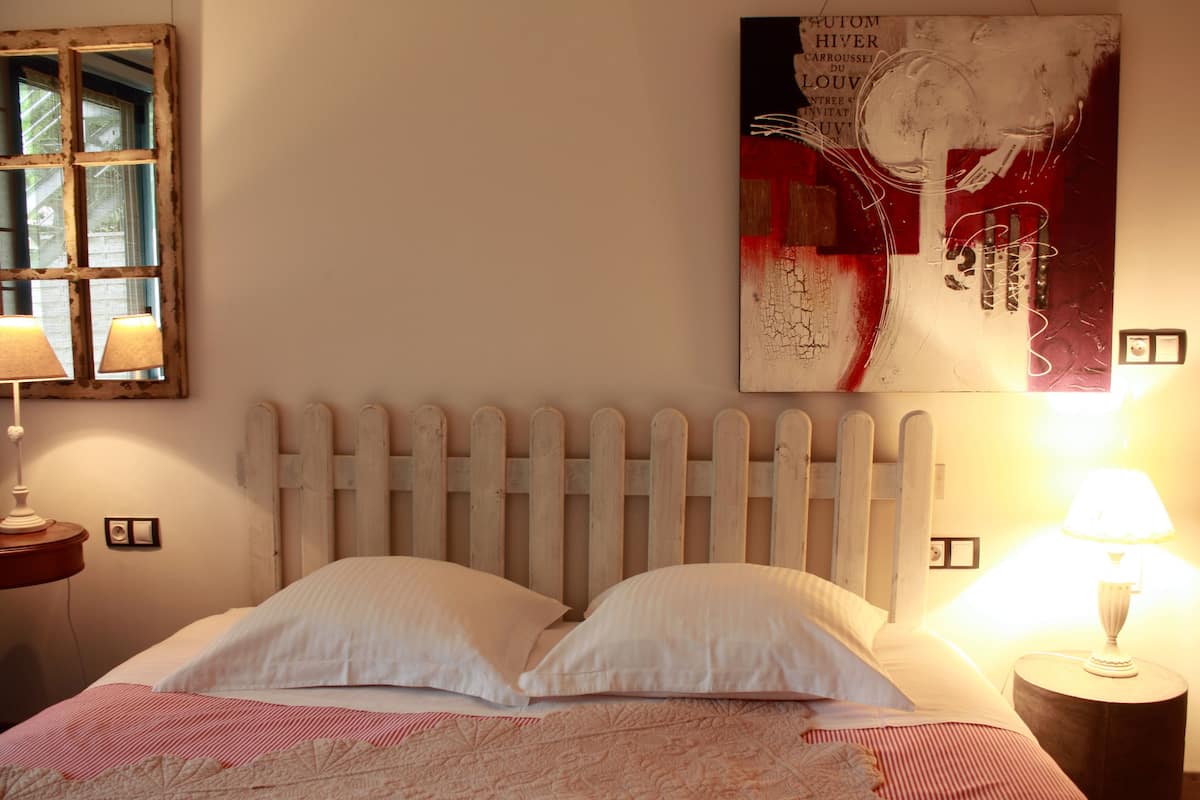 Une chambre à coucher avec une tête de lit en bois, deux lampes de chevet, un lit avec une literie blanche, une fenêtre donnant sur un paysage agréable et un tableau abstrait accroché au mur.