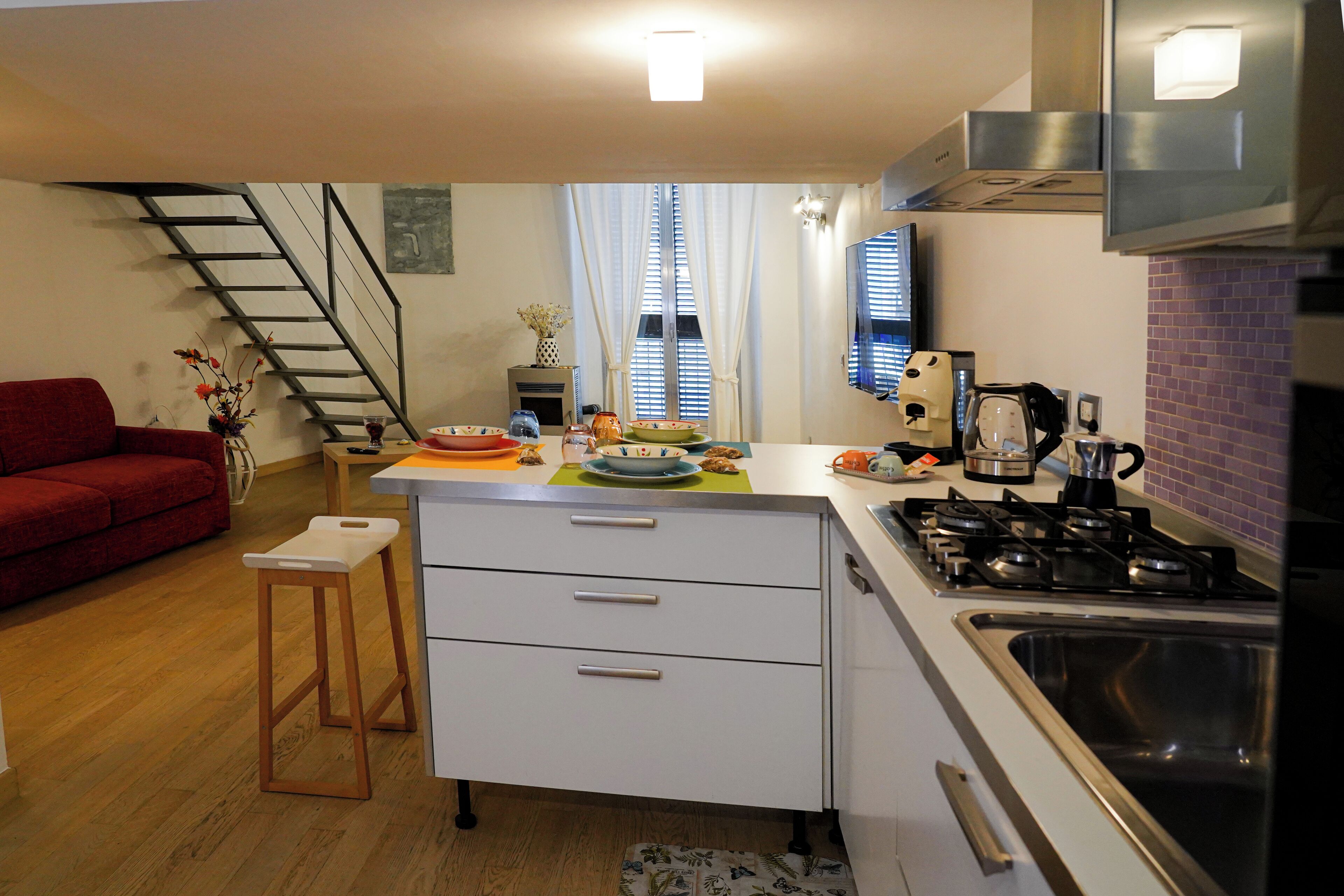 A compact kitchen with a gas stove, a sink, and a small dining area with a table and chairs. A staircase is visible in the background leading to another level.
