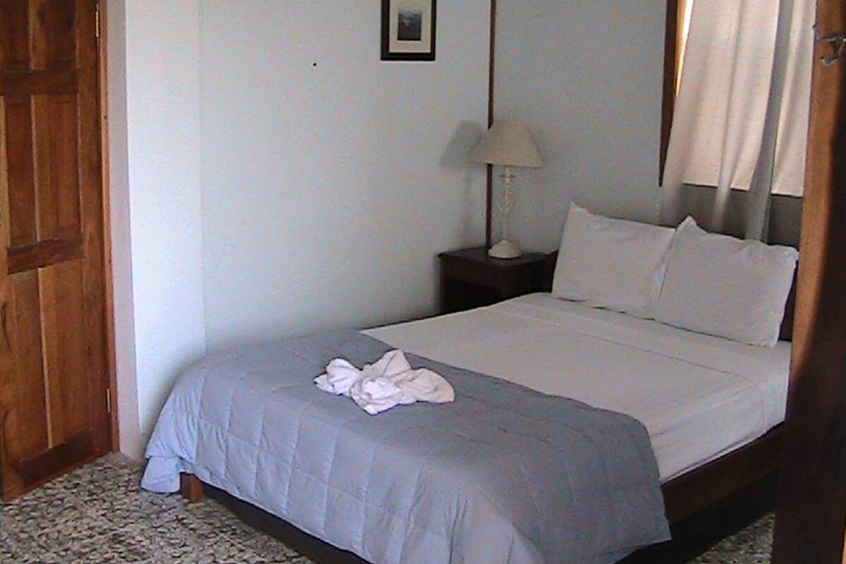 Un dormitorio con cama, mesita de noche, lámpara, puerta y un cuadro en la pared.
