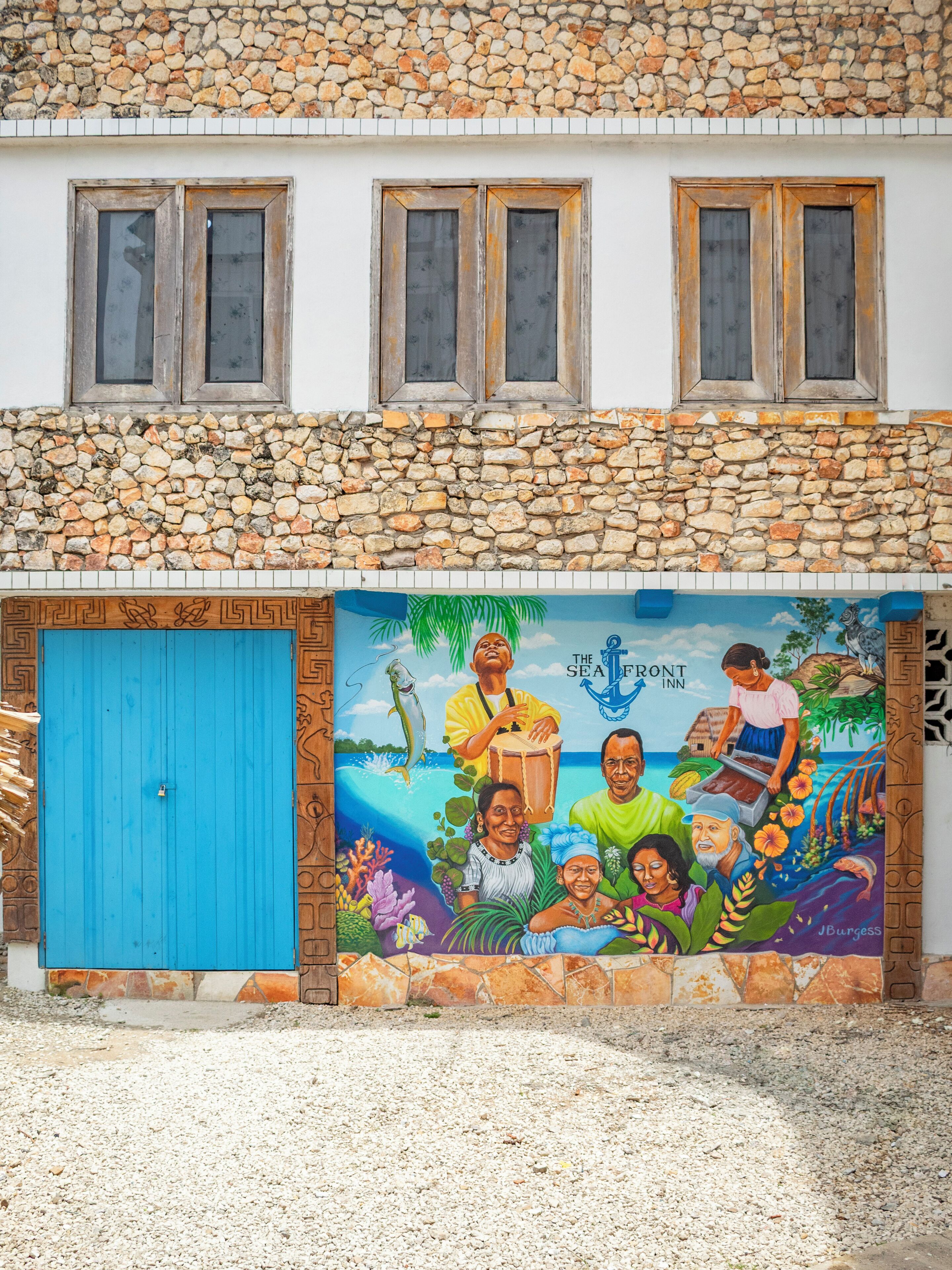 A building with a stone facade, a blue door, and a mural depicting a tropical scene with people and marine life.