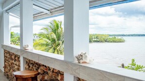 Un balcón con pared de piedra, un banco de madera y vista a un lago con palmeras y una orilla distante.