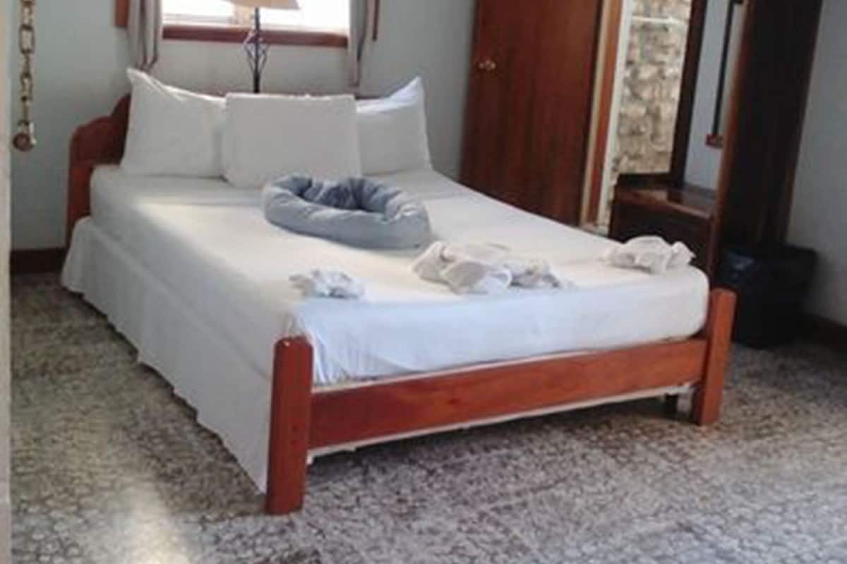 Un dormitorio con cama de madera, ropa de cama blanca, una ventana con cortinas, una lámpara de noche y una pared de piedra.