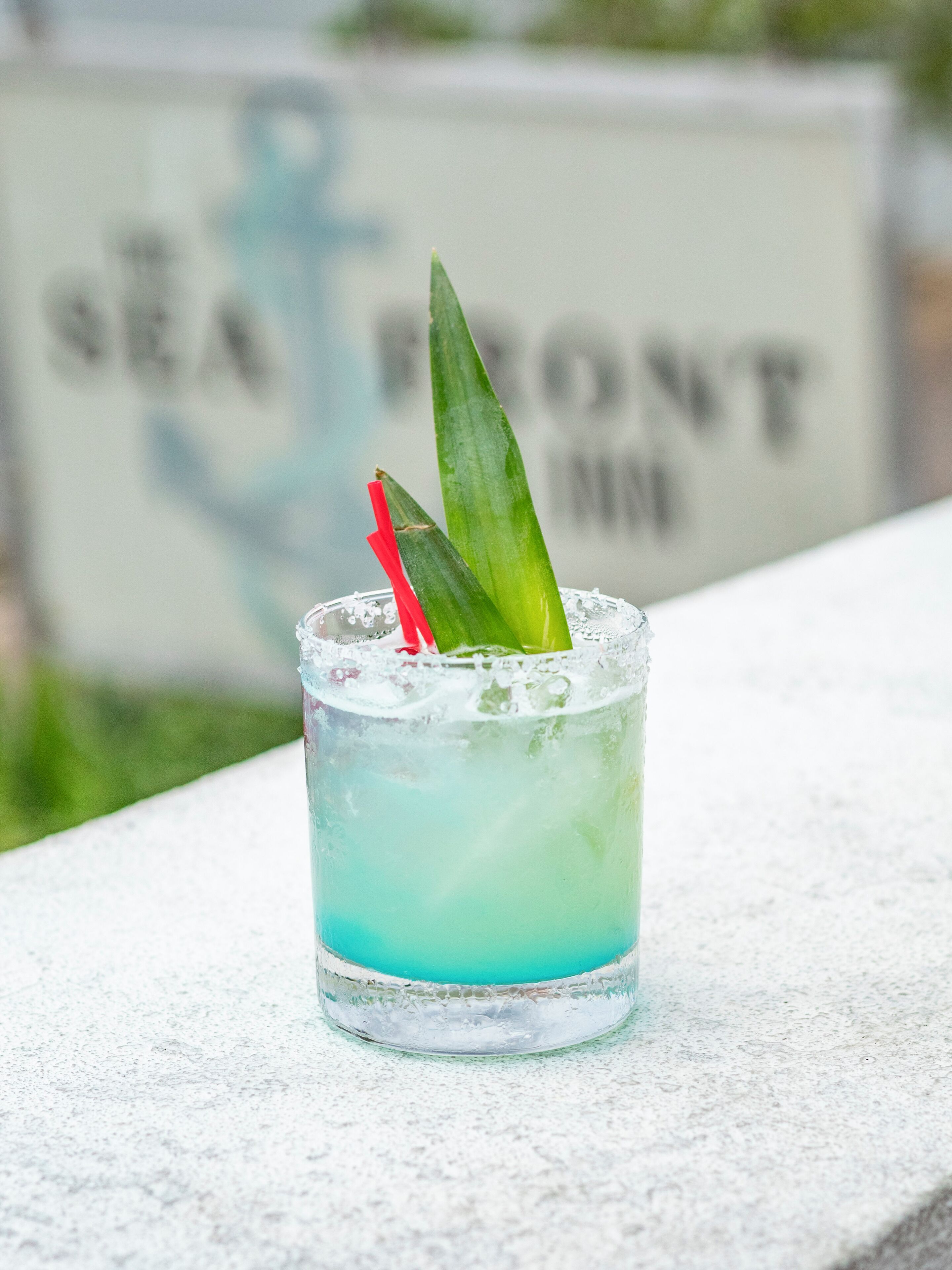 A glass with a blue liquid, garnished with a green leaf and a red straw, placed on a white surface with a blurred background featuring a sign with text.