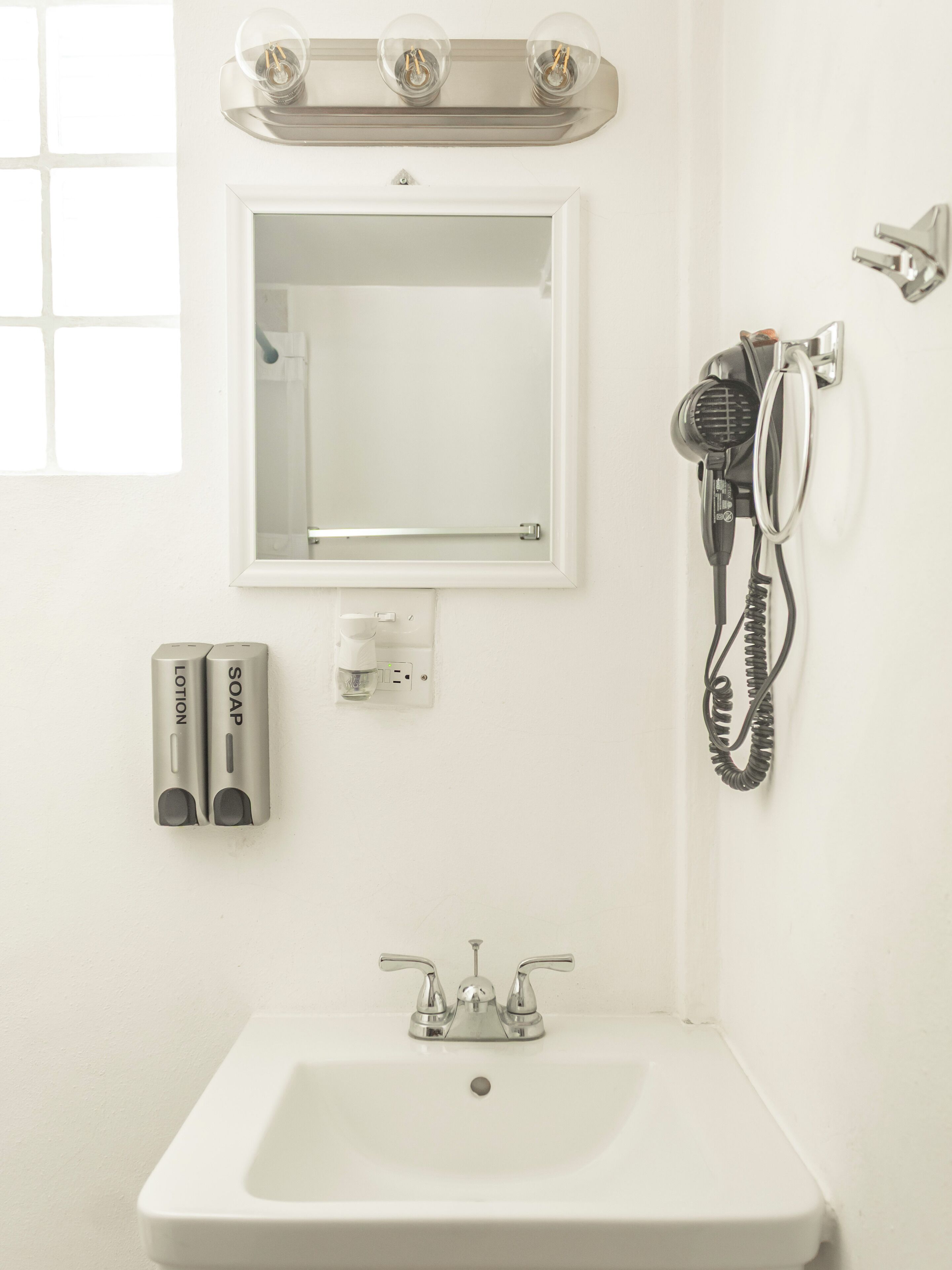 Une salle de bain avec un lavabo blanc, un miroir, un sèche-cheveux et deux distributeurs de savon.