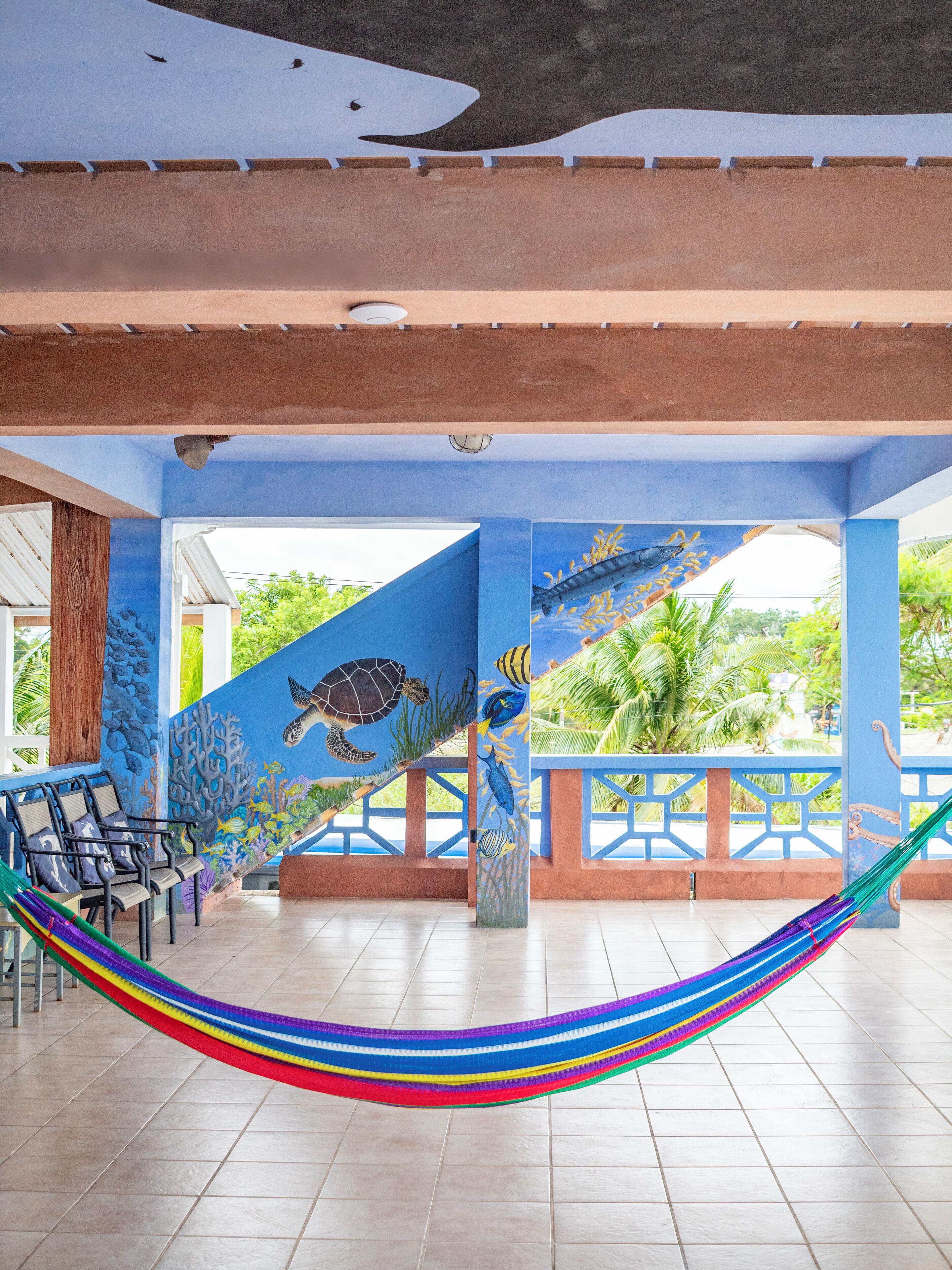 A covered outdoor seating area with a colorful hammock, blue walls with marine life murals, and a view of greenery.