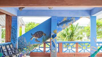 A covered outdoor seating area with a colorful hammock, blue walls with marine life murals, and a view of greenery.