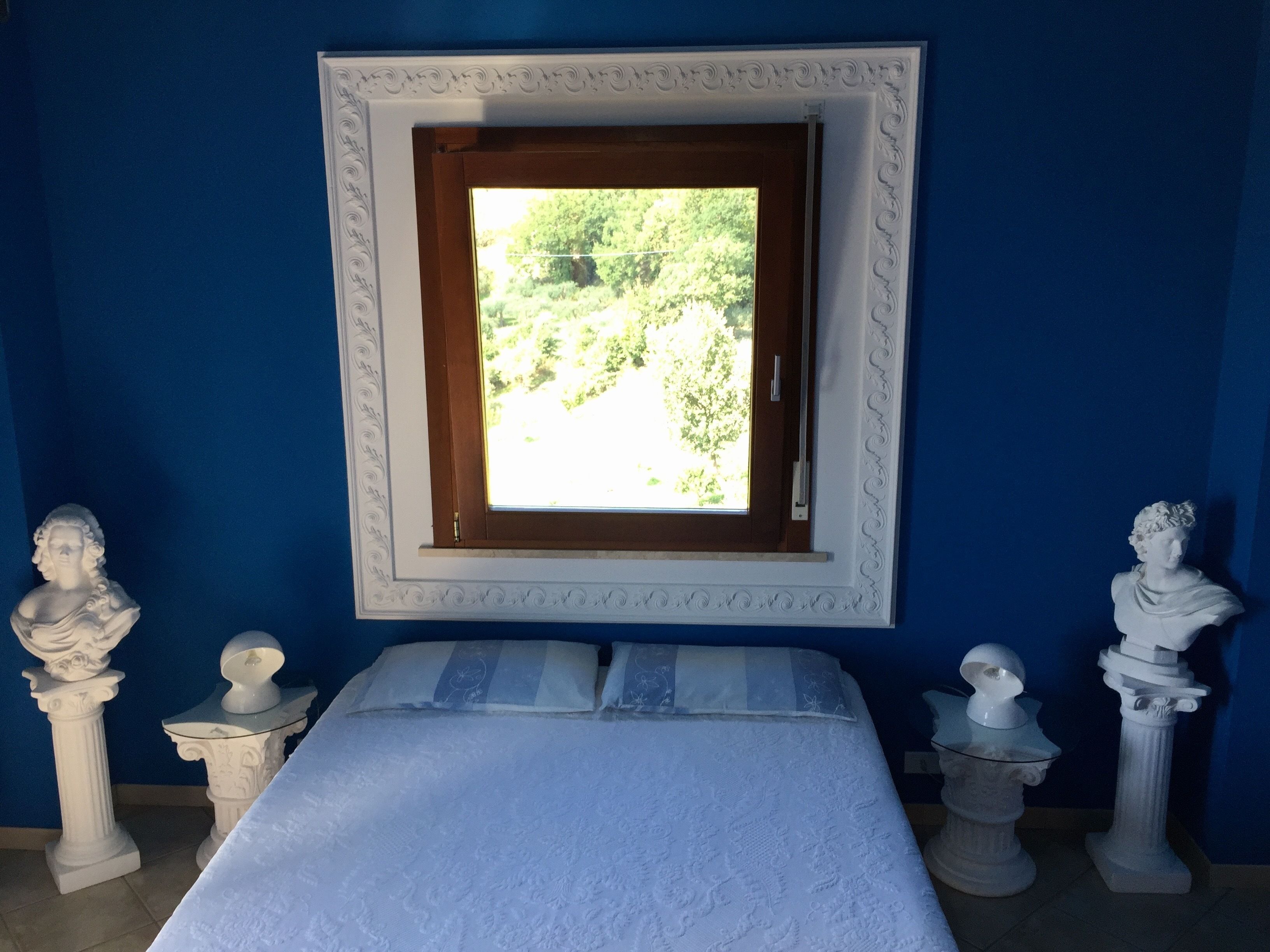 A room with a bed, a window with a view of greenery, and two bust sculptures on pedestals.