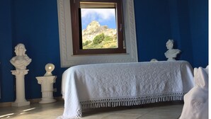 Una habitación con cama, ventana con vistas a una montaña y esculturas clásicas.