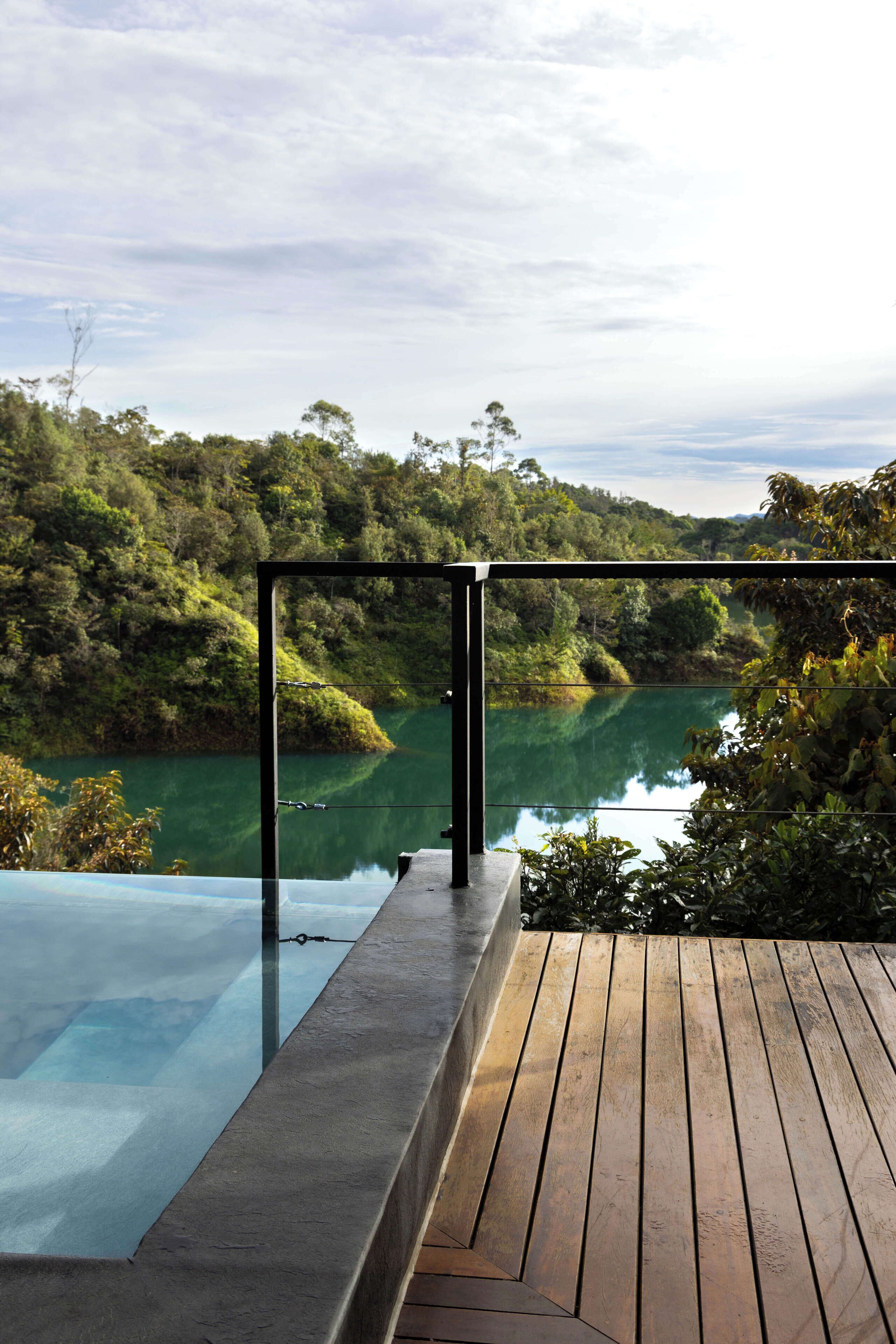 Piscina infinita com deck de madeira e guarda-corpo de vidro, com vista para um rio e colinas arborizadas.
