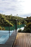 Piscina infinita con terraza de madera y barandilla de cristal que da a un río y colinas boscosas.