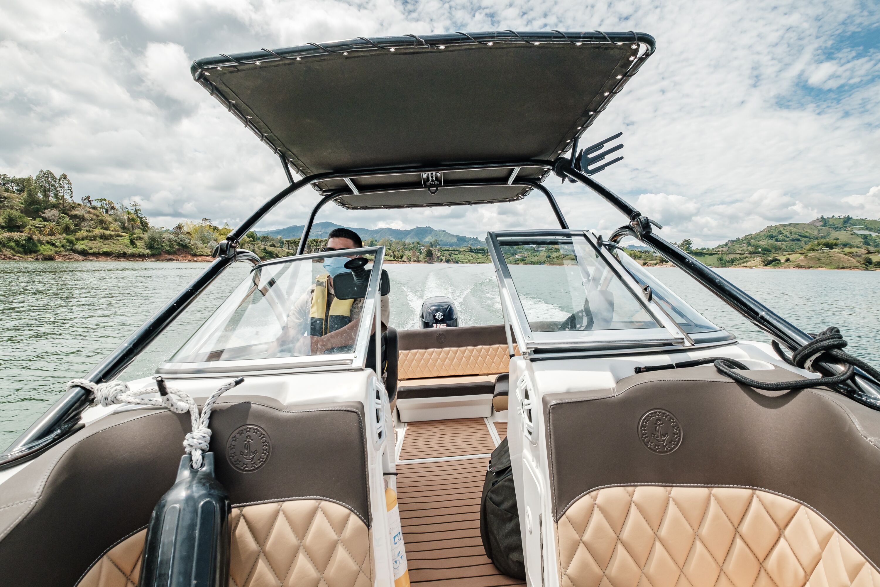 A boat interior with a cushioned seating area, a steering wheel, and a view of a river with greenery in the background.