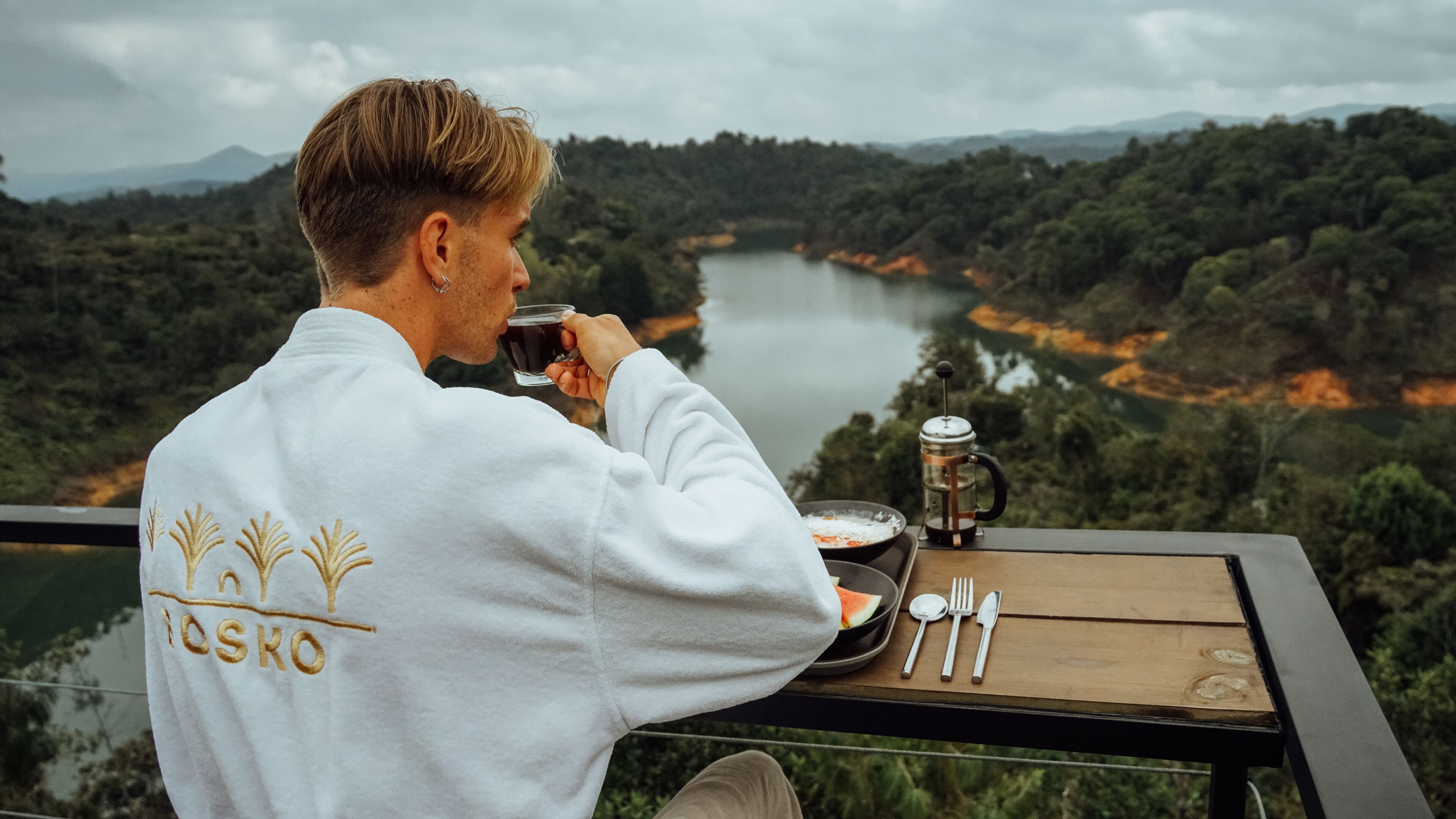 Una persona sentada a una mesa con vista a un lago y un bosque, sosteniendo una taza. La mesa tiene una cafetera, un plato con comida y cubiertos. La camisa de la persona tiene un logotipo.