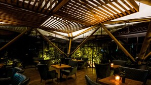 An indoor dining area with wooden tables and chairs, a sloped ceiling with wooden beams, and a view of greenery outside. - Bosko (Guatapé)