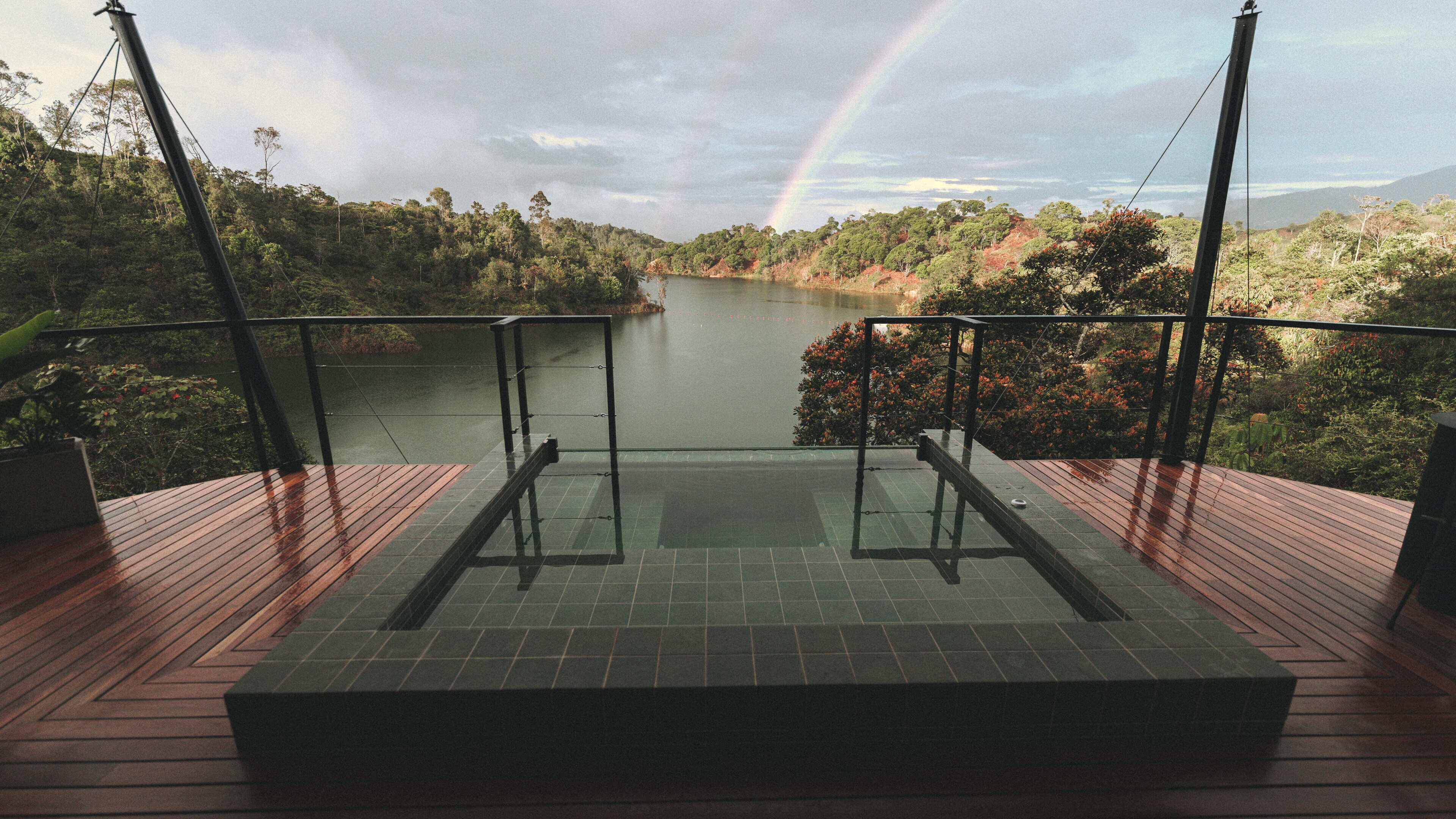 Una terraza con barandilla de vidrio que da a un lago y un arcoíris.
