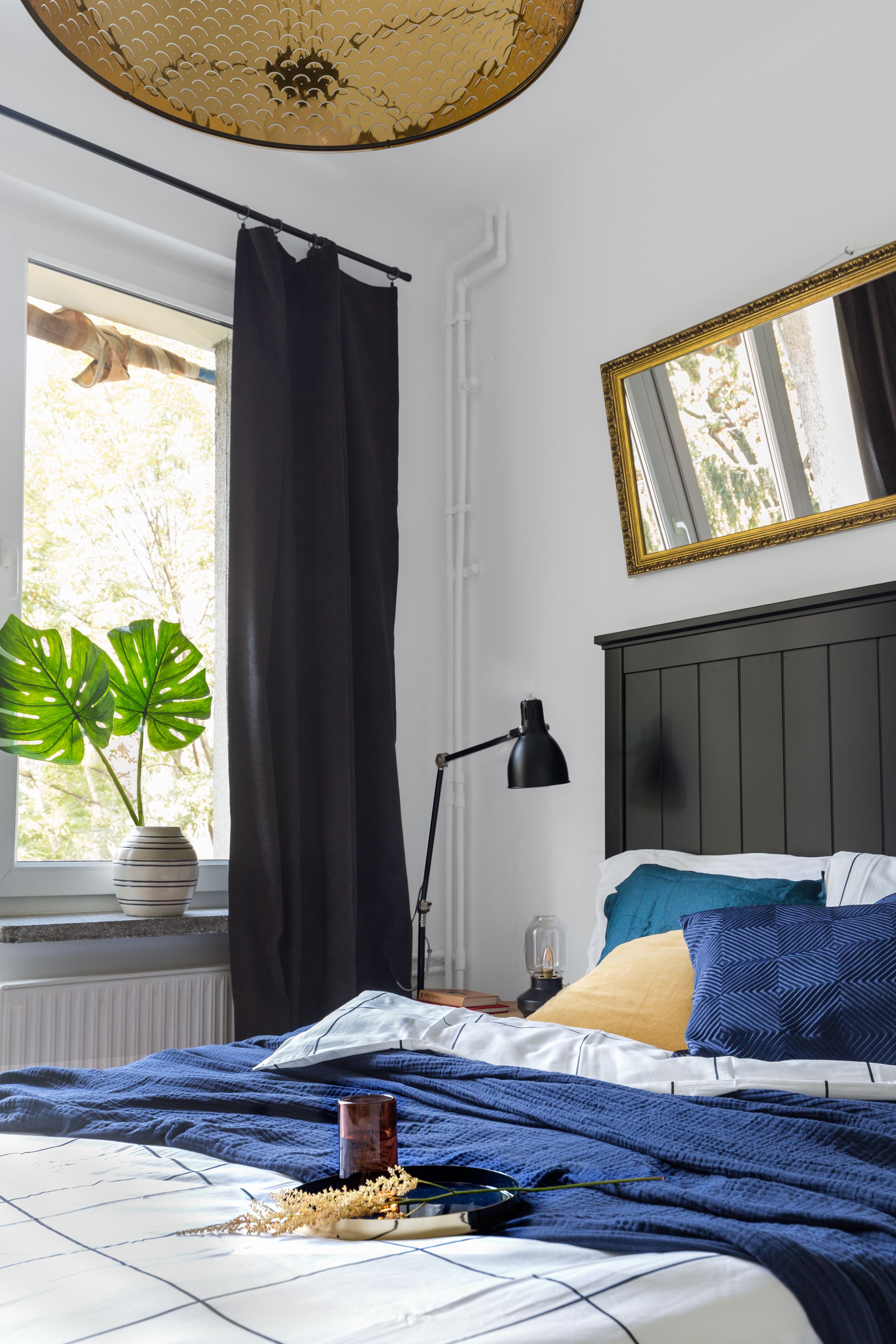 A bedroom with a bed, a window with curtains, a potted plant, and a mirror on the wall.