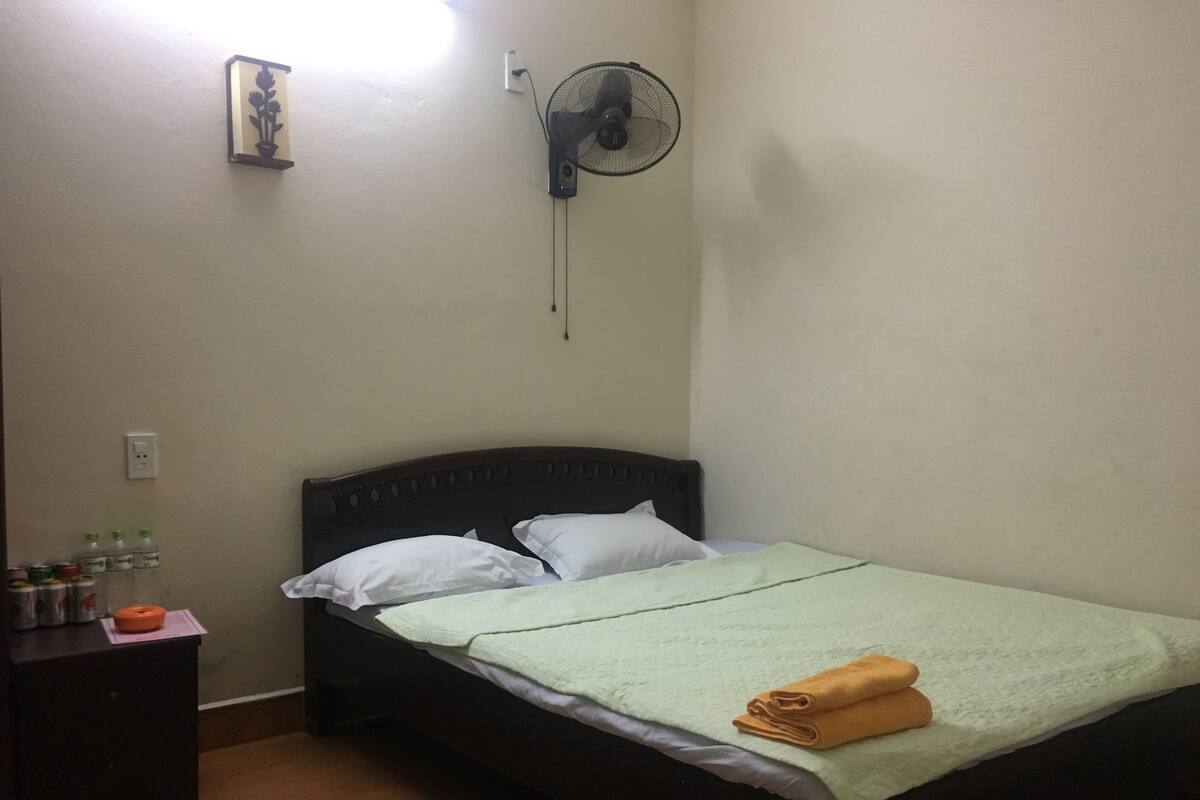 Ein Einzelbett mit grüner Decke und zwei Kissen, ein Nachttisch mit Lampe sowie ein an der Wand befestigter Ventilator.