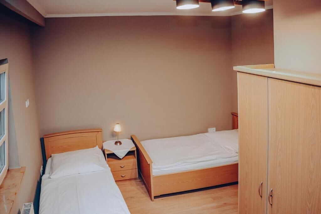 A bedroom with two single beds, a wooden wardrobe, a nightstand with a lamp, and a window.